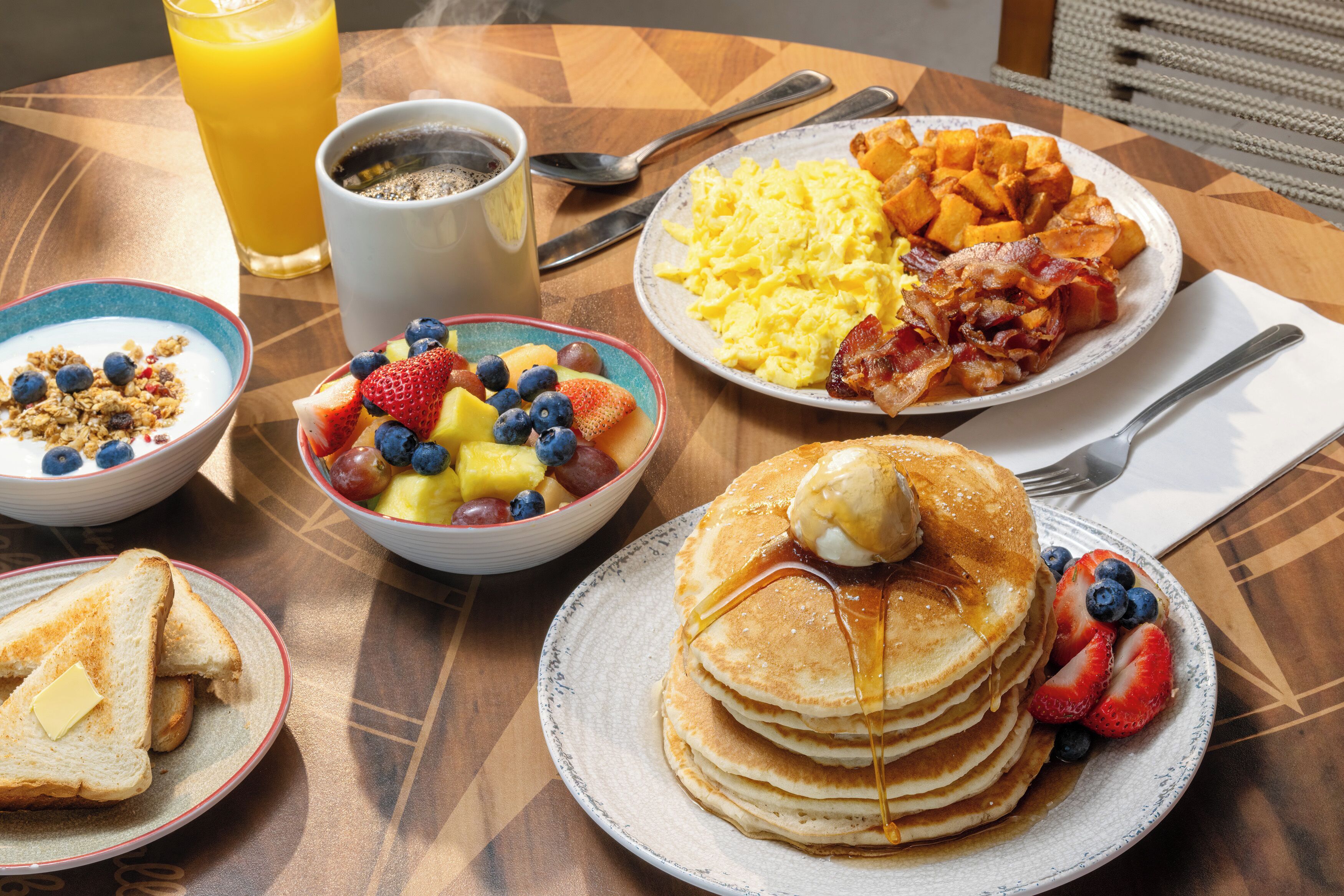 Una mesa de desayuno con panqueques, huevos revueltos, tocino, tostadas, fruta y una taza de café.