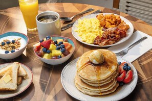 Una mesa de desayuno con panqueques, huevos revueltos, tocino, tostadas, fruta y una taza de café.