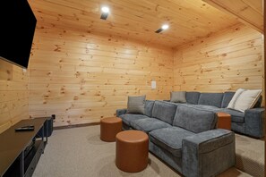A cozy room with wooden walls, a grey sectional sofa, two brown ottomans, and a black TV stand. - Frog Hill | Hocking Hills, EV Charger, Hot Tub (Logan)