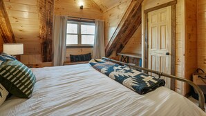 A wooden cabin bedroom with a bed, a window with curtains, a door, and a staircase. - Frog Hill | Hocking Hills, EV Charger, Hot Tub (Logan)