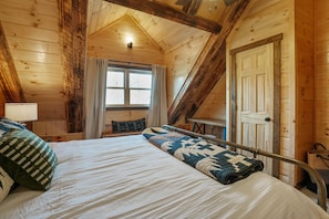A wooden cabin bedroom with a bed, a window with curtains, a door, and a staircase. - Frog Hill | Hocking Hills, EV Charger, Hot Tub (Logan)