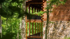 A log cabin with a stone wall and a wooden staircase leading to a balcony. - Frog Hill | Hocking Hills, EV Charger, Hot Tub (Logan)