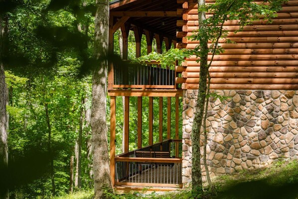 A log cabin with a stone wall and a wooden staircase leading to a balcony. - Frog Hill | Hocking Hills, EV Charger, Hot Tub (Logan)