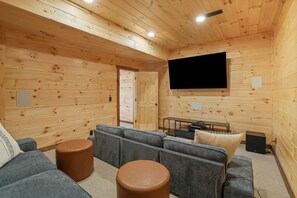 Interior acolhedor de uma cabana de madeira, com uma televisão grande, sofás cinza e uma pequena mesa.