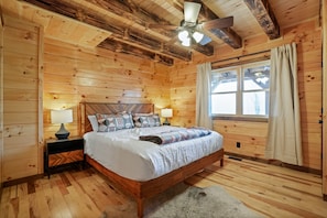A wooden cabin bedroom with a bed, two bedside tables, a ceiling fan, and a window with curtains. - Frog Hill | Hocking Hills, EV Charger, Hot Tub (Logan)