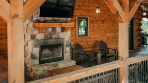 A log cabin interior with a stone fireplace, a mounted television, and two Adirondack chairs. - Frog Hill | Hocking Hills, EV Charger, Hot Tub (Logan)