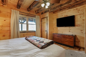 Un dormitorio de cabaña de madera con una cama, un televisor montado en la pared y una ventana con cortinas.