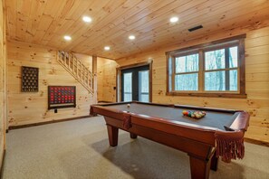 A wooden room with a pool table, a staircase, and a window with a view of trees.