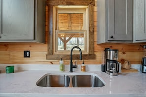 A kitchen with a double sink, wooden wall paneling, a window with a view, and a coffee maker.