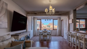 A spacious living area with a large flat-screen TV, a dining table with chairs, a sofa with cushions, and a kitchen area with a bar.