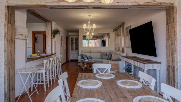 A dining area with a wooden table, white chairs, and a chandelier. There is a bar area with stools and a television mounted on the wall.