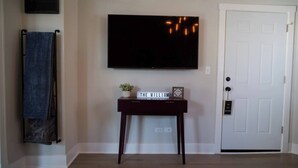 Una habitación con un televisor de pantalla plana montado en la pared, una consola de madera oscura con una planta pequeña y un letrero que dice "THE WILLIAM", un perchero con una toalla azul y una puerta blanca con una llave en la cerradura.