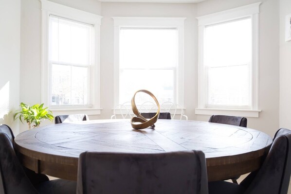 Une table ronde ornée d’un centre de table original, entourée de chaises, dans une pièce dotée de grandes fenêtres et agrémentée d’une plante en pot.