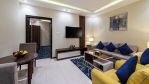 A modern hotel room with a flat-screen TV, a sofa with blue and yellow cushions, a dining table with fruit, and a patterned rug. - Al Qasr Hotel Suites -Al Masyaf District (Khamis Mushait)