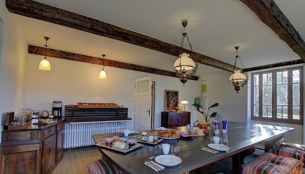 A dining area with a long table, wooden beams, pendant lights, and a view through large windows. - Goute la vie  (Marchamp)
