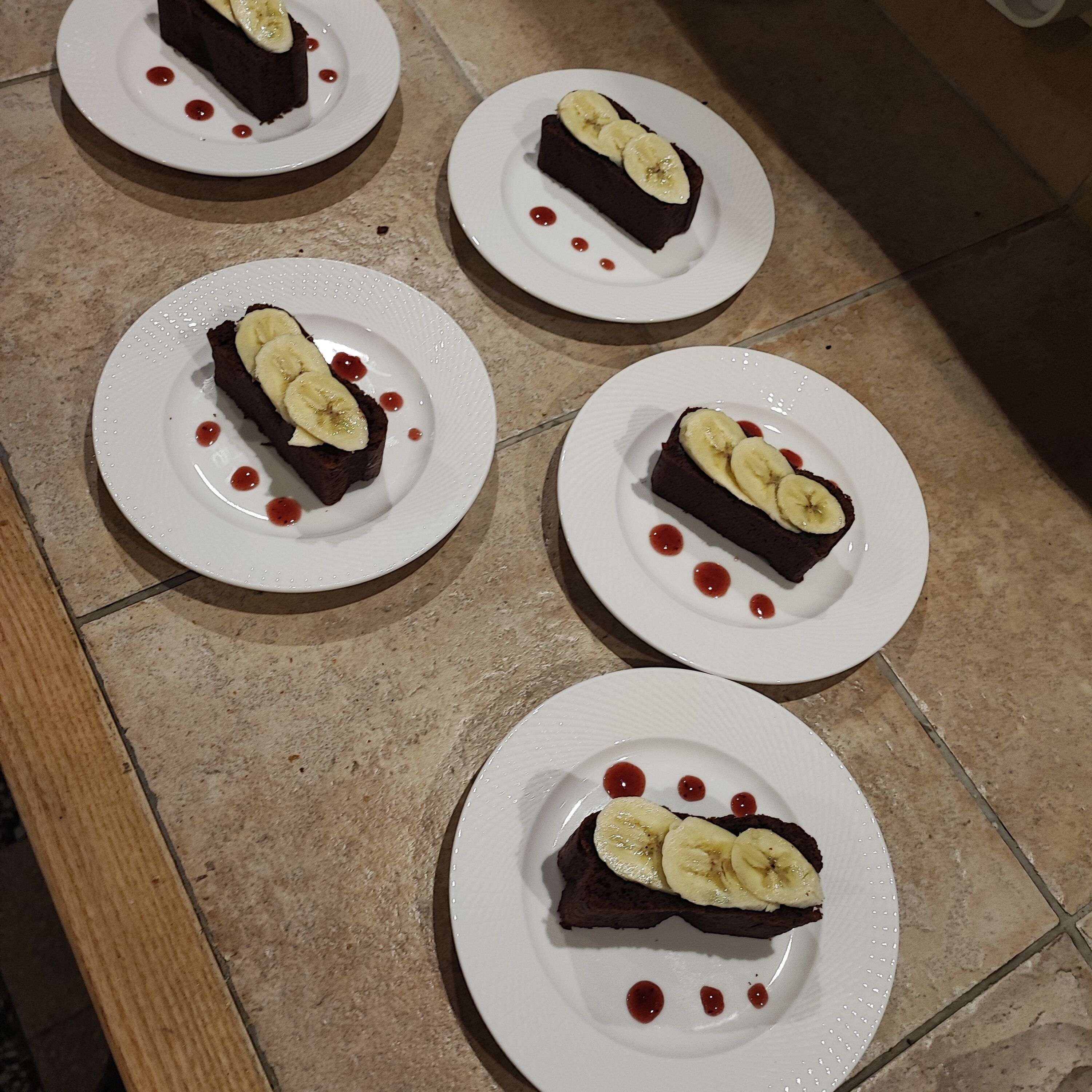 Four plates with chocolate cake slices topped with banana slices and red sauce.