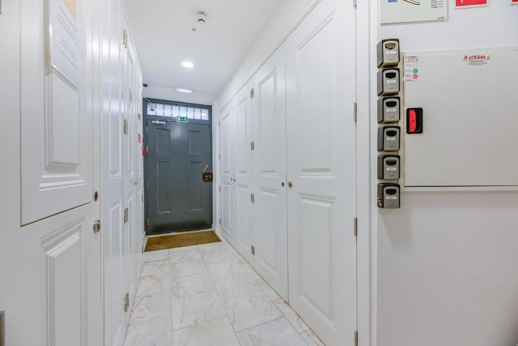 A hotel corridor with white doors, a grey door at the end, and a wall-mounted elevator control panel.