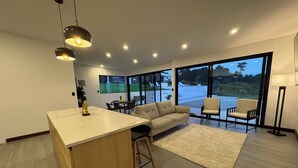 A modern kitchen with a central island, a dining area with a table and chairs, a sofa, and a large window offering a view of the outdoors.