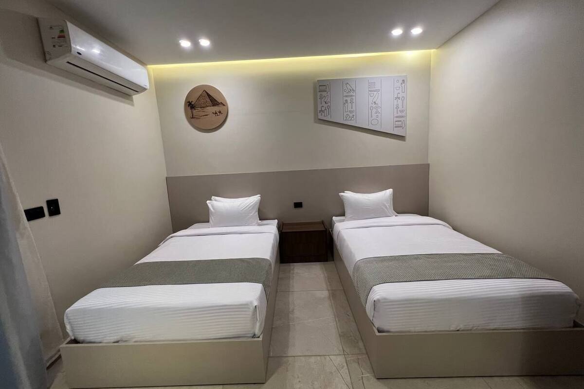 A hotel room with two single beds, an air conditioning unit, a wall-mounted light fixture, and a small table.