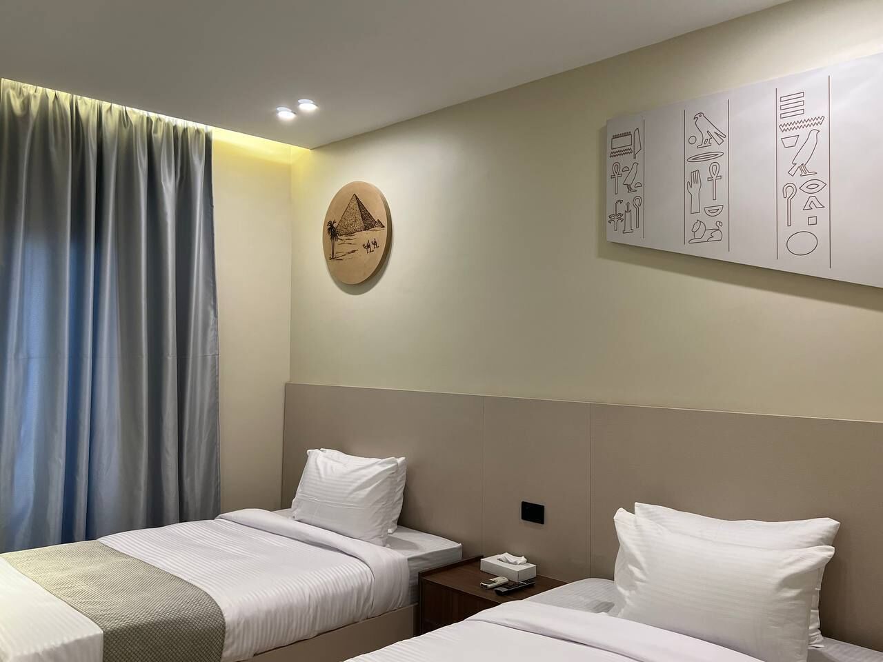 Habitación de hotel con dos camas, una mesita de noche, un reloj de pared y una pared con arte decorativo.