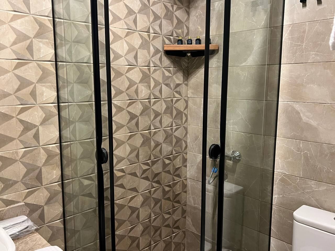 A shower enclosure with geometric patterned tiles, a glass door, and a wooden shelf with toiletries.