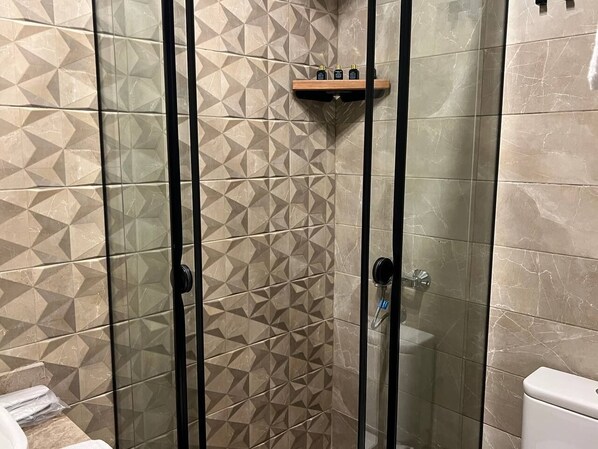 Un espace de douche avec des carreaux aux motifs géométriques, une porte en verre et une étagère en bois où sont rangés les articles de toilette.
