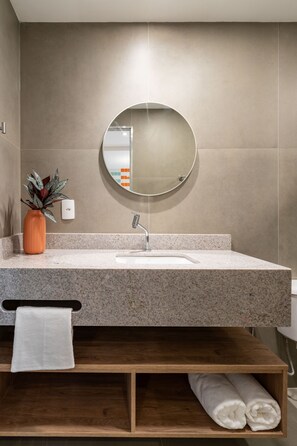 A modern bathroom with a granite countertop, a round mirror, a wooden shelf, and a potted plant.