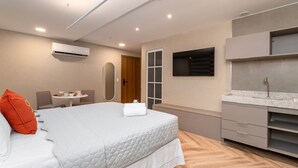 A modern hotel room with a large bed, a built-in TV, a small dining area, and a bathroom with a modern sink and mirror.