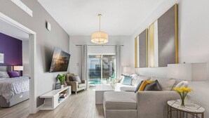 A modern living room with a sofa, a coffee table, and a TV. There is a window with a view of the outside. - Serenity Townhouse! Private Pool and Pet-Friendly (Davenport)