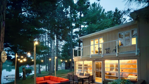 A two-story house with a patio area featuring a sofa and dining table. - Beautiful Lakefront cabin woodburning fireplace, grill, canoe, SUP, AC, parking. (Wautoma)