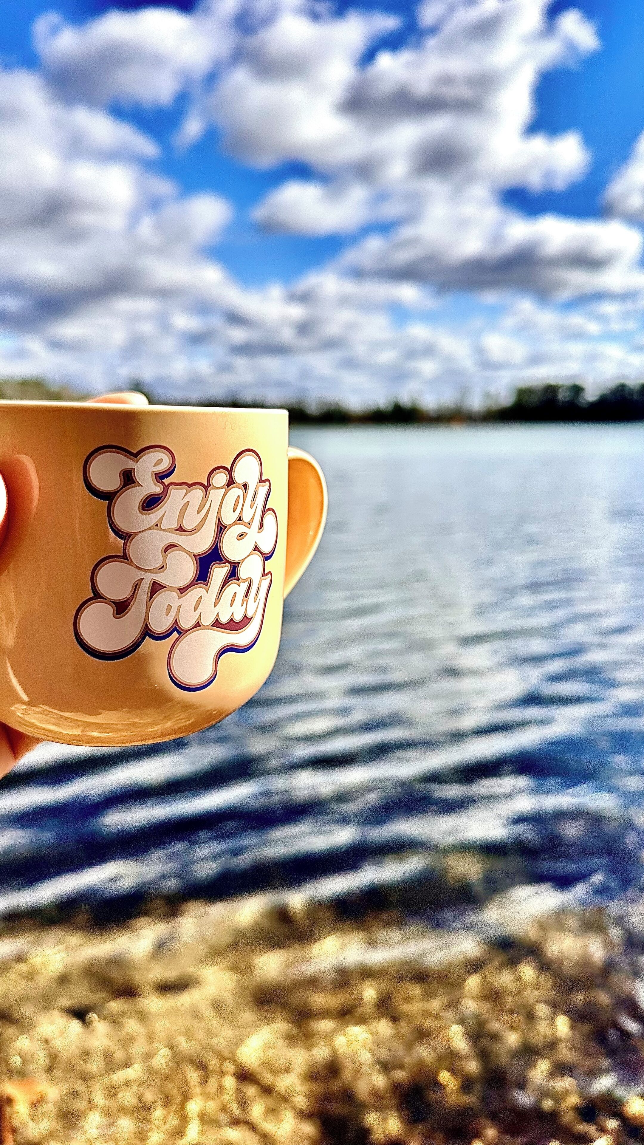 Une main tenant une tasse sur laquelle est inscrit le message « Passez une bonne journée », au-dessus d’une étendue d’eau sous un ciel dégagé.