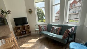 A modern living room with a sofa, a television, a wooden floor, and large windows. - Sea View – Stunning Three‑Bedroom Apartment with Panoramic Coastal Views (Eastbourne)