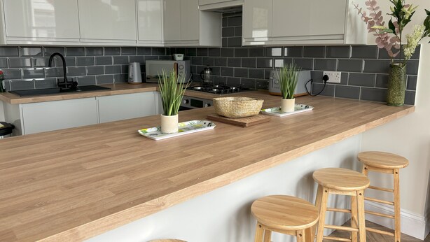 A modern kitchen with white cabinets, a wooden countertop, and a built-in stove. There are three stools at the counter and a decorative plant on the wall. - Sea View – Stunning Three‑Bedroom Apartment with Panoramic Coastal Views (Eastbourne)