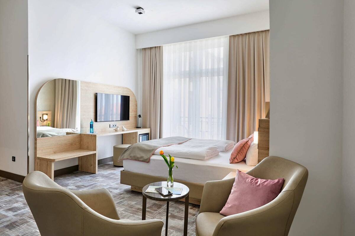 Een hotelkamer met een bed, een bureau met een televisie, een stoel en een tafeltje met een bloemenvaas.