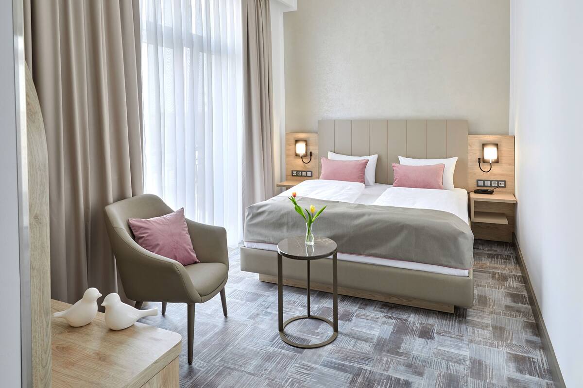 Een hotelkamer met een bed, twee nachtkastjes met lampen, een stoel, een klein tafeltje met een vaas en een raam met gordijnen.