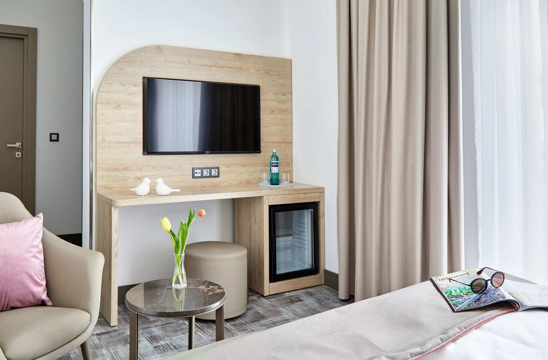 A hotel room with a TV mounted on a wooden wall, a minibar, a small table with a vase of flowers, a bed with a magazine and glasses, and a window with curtains.