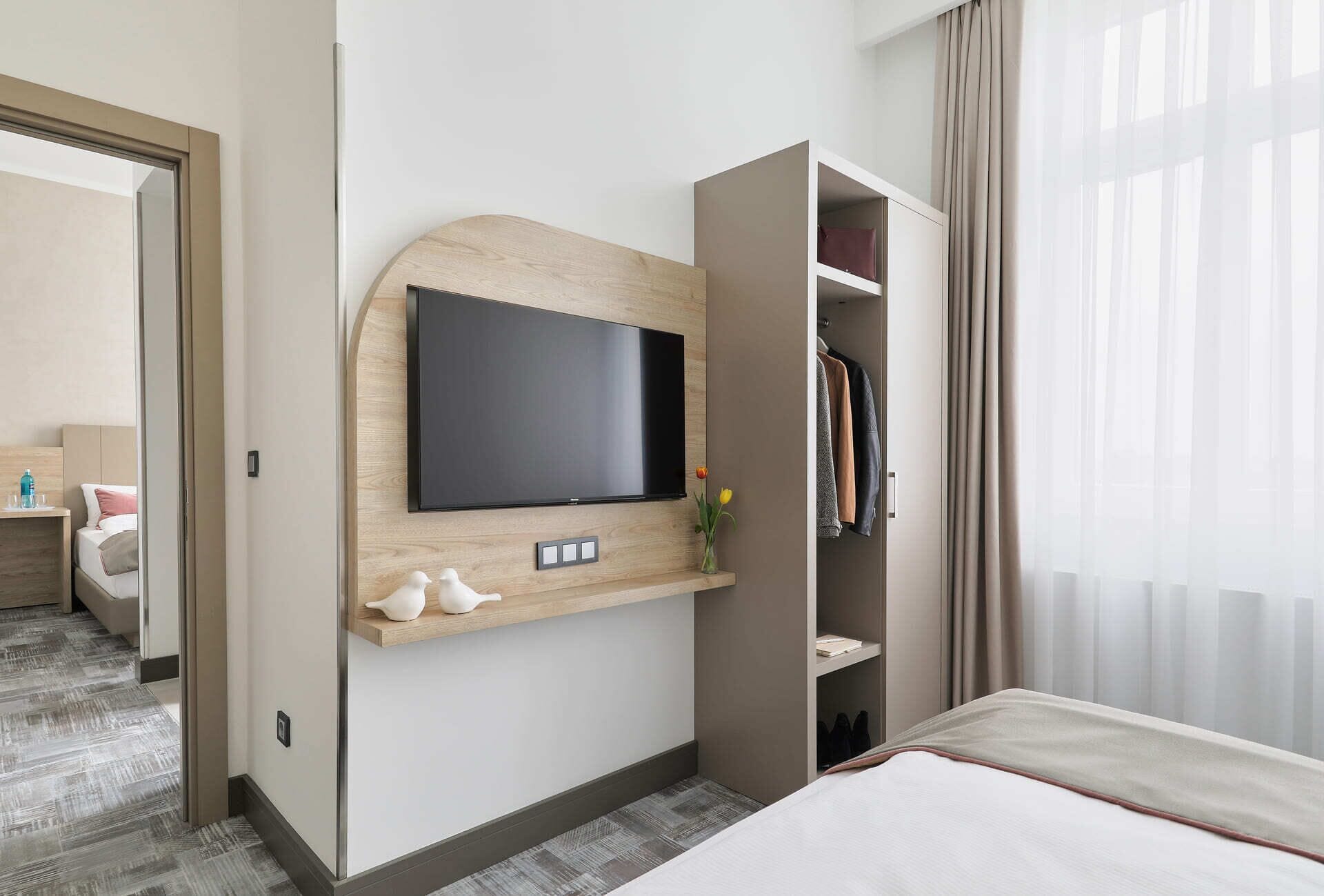 Une chambre d’hôtel équipée d’un téléviseur fixé au mur, d’une étagère en bois agrémentée d’objets décoratifs, d’une armoire et d’un lit.