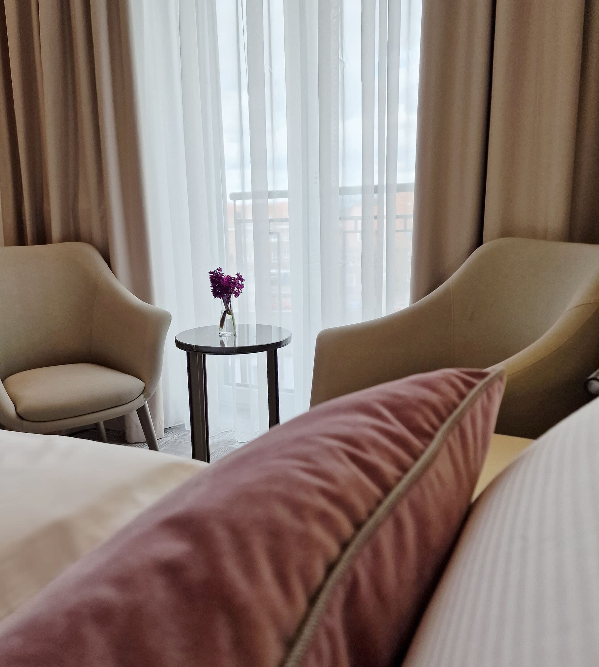 Una habitación de hotel con dos sillones, una mesita redonda con un jarrón de flores moradas y una cama con almohada rosa.