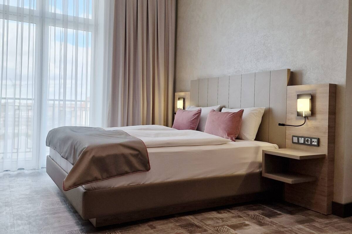 Een moderne hotelkamer met een groot bed, een nachtkastje met een lamp en uitzicht op de stad door grote ramen.