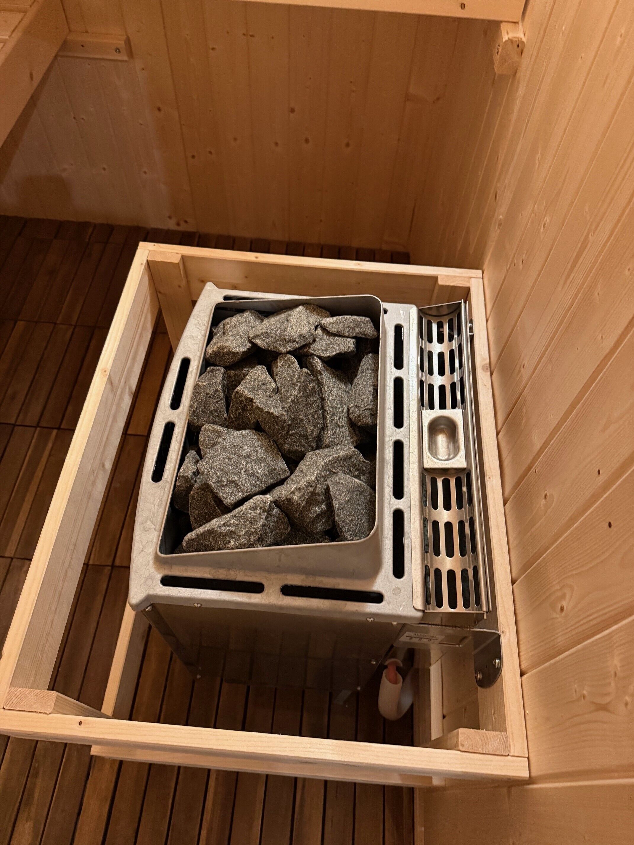 Uma sauna com interior em madeira, uma grade de metal cheia de pedras e um pequeno recipiente metálico.