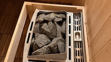 Uma sauna com interior em madeira, uma grade de metal cheia de pedras e um pequeno recipiente metálico.