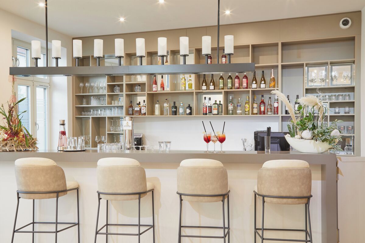 A modern bar area with a long counter, multiple shelves of bottles, glasses, and a decorative plant arrangement.