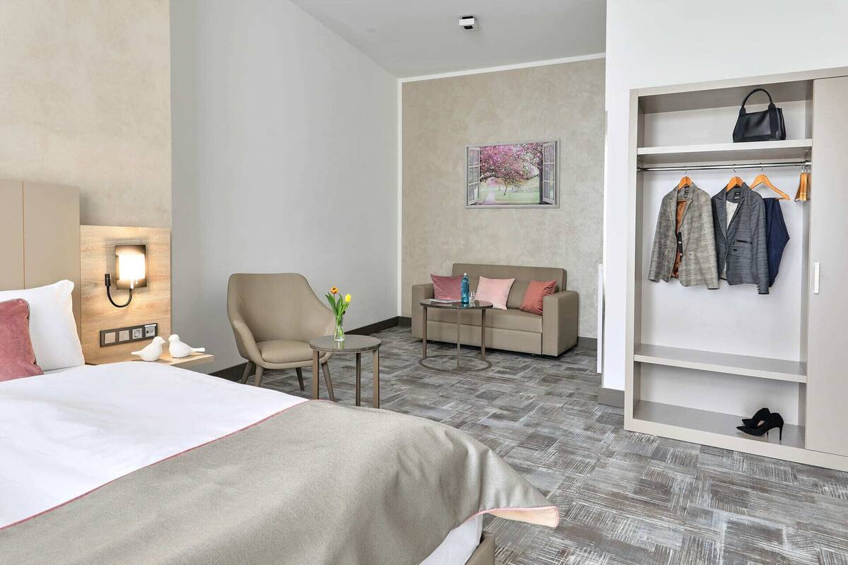 Een hotelkamer met een bed, een slaapbank, een stoel, een kleine tafel met een vaas, en een kledingkast met kleding en schoenen.