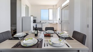Un espace repas moderne avec une longue table dressée pour six personnes, une cuisine en arrière-plan et une grande fenêtre laissant entrer la lumière naturelle.