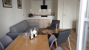 Uma sala de estar moderna com um sofá cinzento, uma mesa de jantar de madeira com copos e garrafas de vinho, uma poltrona cinzenta e uma cozinha ao fundo.