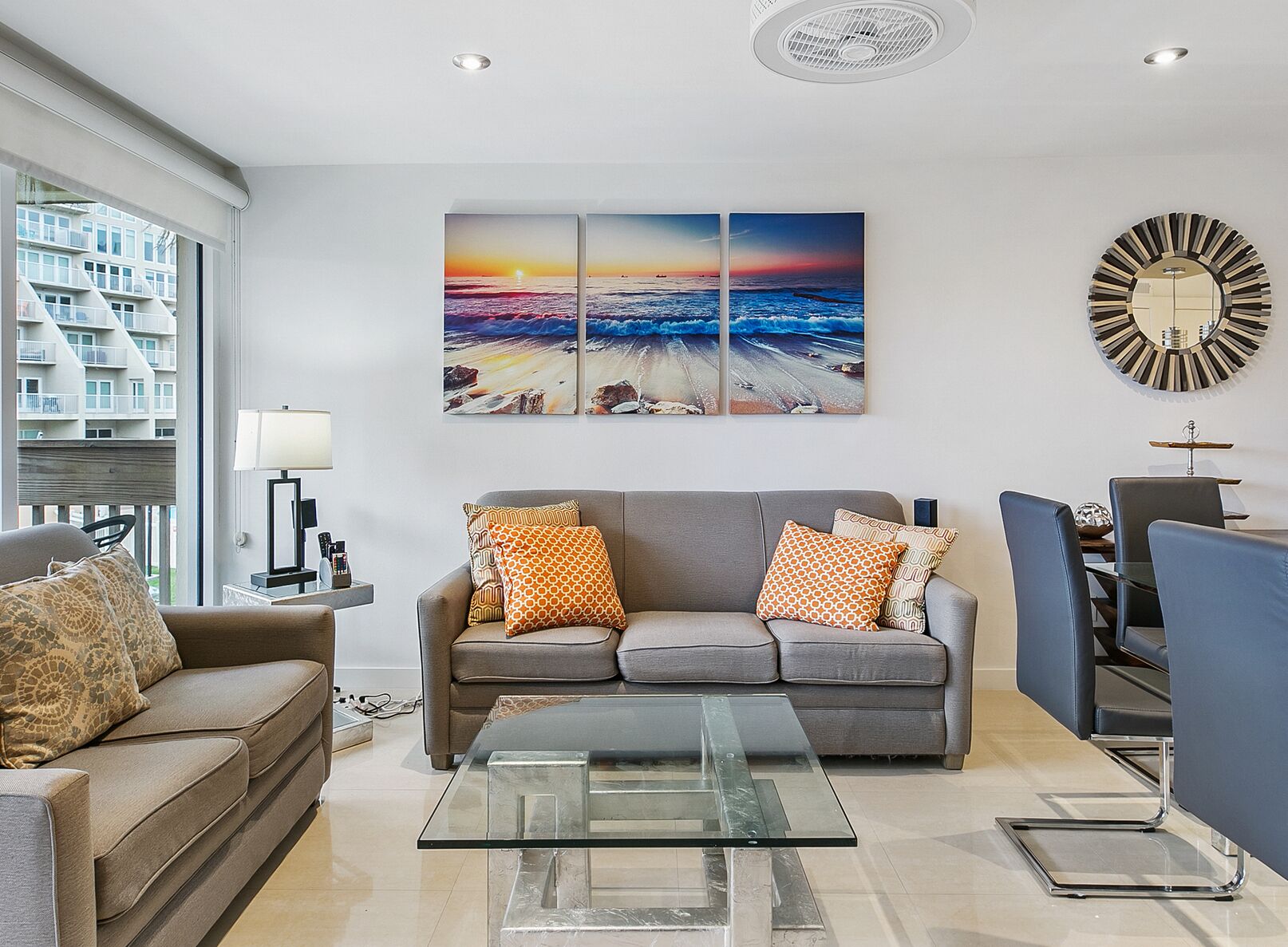 A modern living room with a grey sofa, a glass coffee table, and a large painting of a beach sunset.