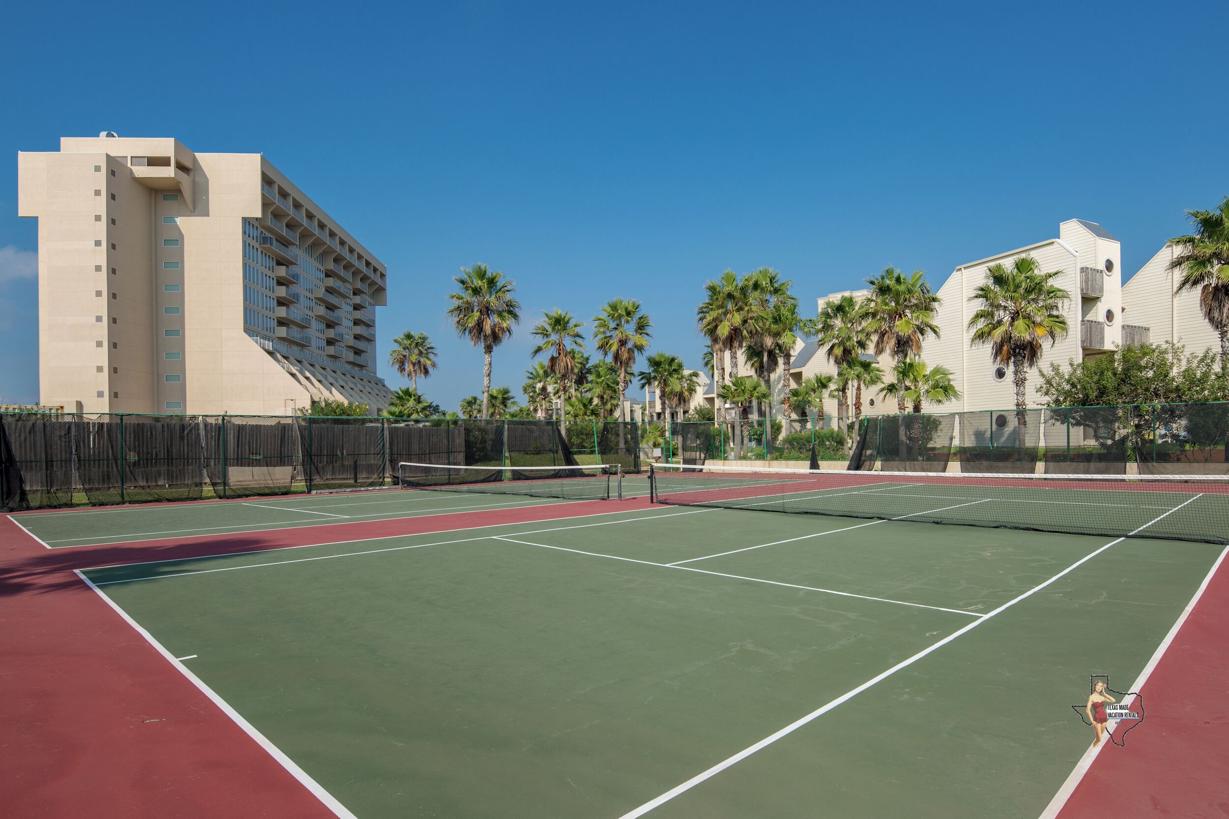 A tennis court with a net, surrounded by palm trees and a building.