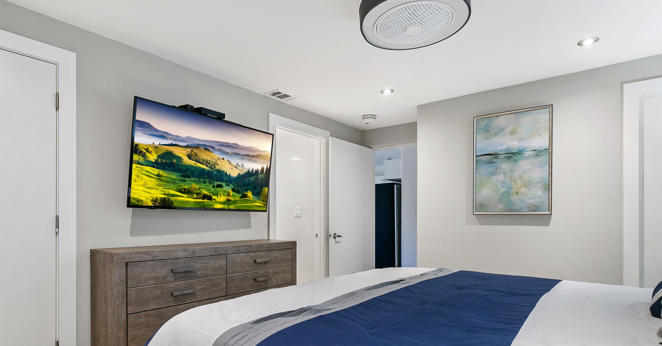 A bedroom with a bed, a dresser, a flat-screen TV mounted on the wall, and a door leading to another room.