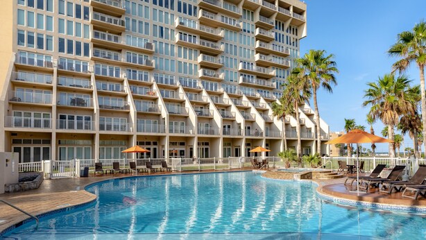 Una piscina ampia circondata da lettini e ombrelloni, sullo sfondo palme e un cielo azzurro terso.
