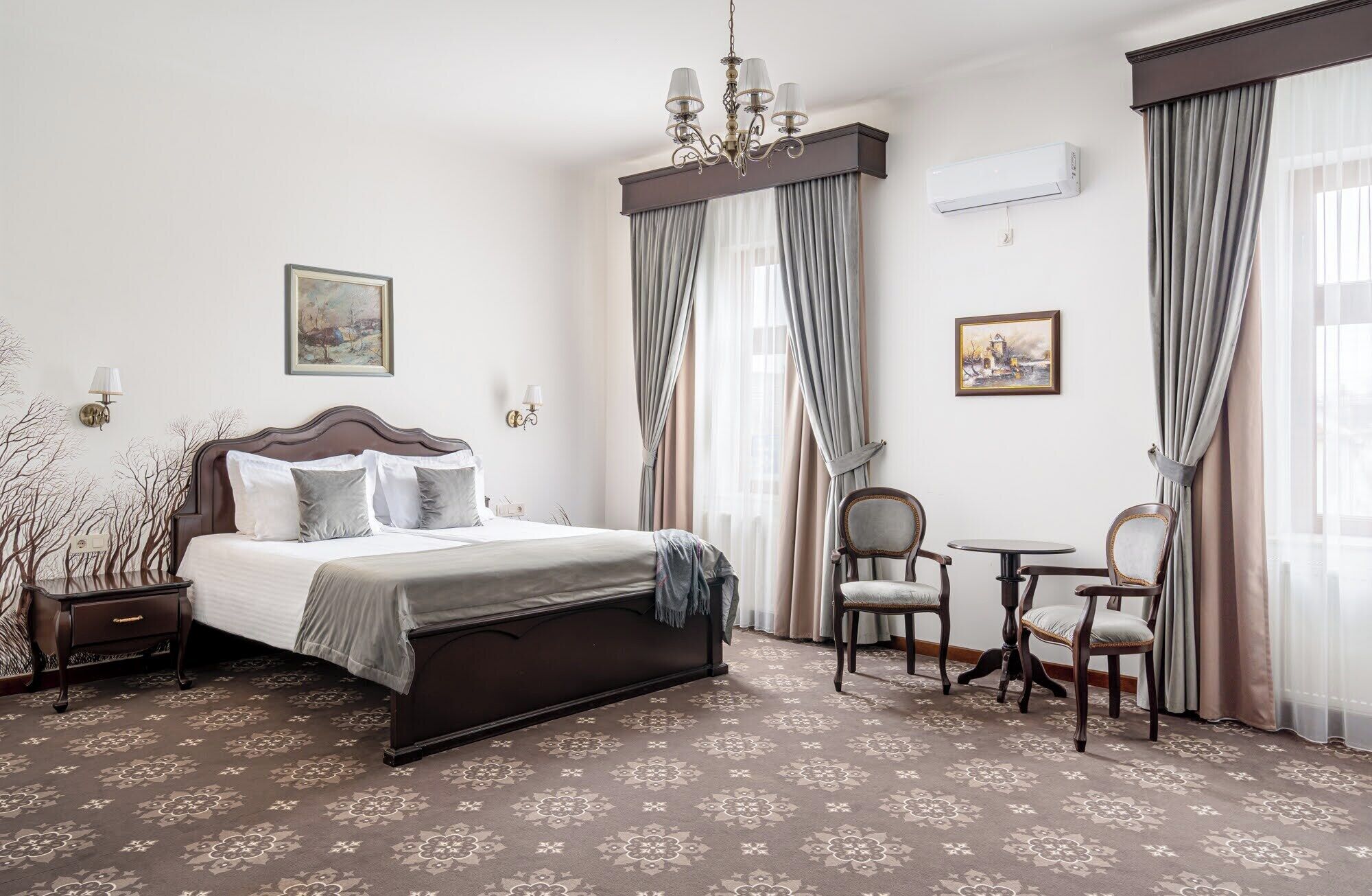 A hotel room with a large bed, a small table with two chairs, a chandelier, and two paintings on the walls.